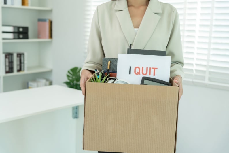 Woman carrying box of office contents and letter reading I QUIT